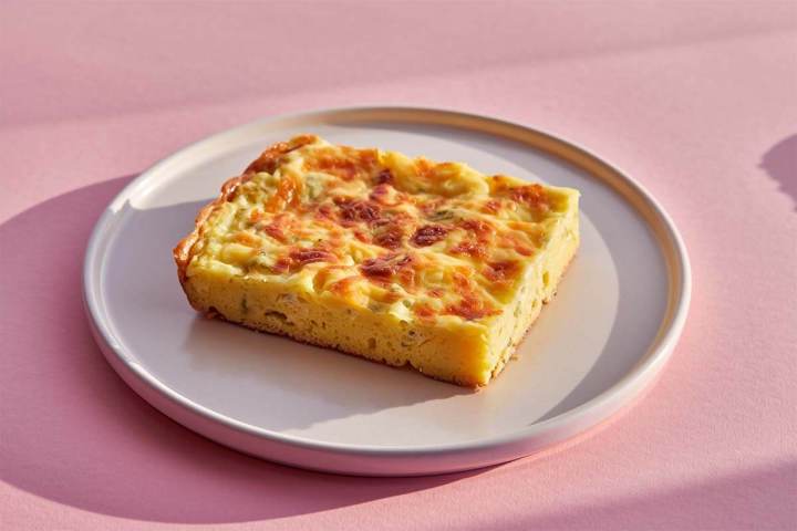 Slice of egg and cheese frittata on a white plate, set on a pink background.