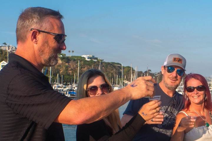 Four people on a boat, toasting with drinks, smiling in Dana Point Harbor with boats in the background.