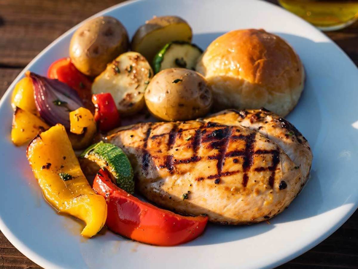 Grilled chicken with vegetables, potatoes, and a bun on a white plate.