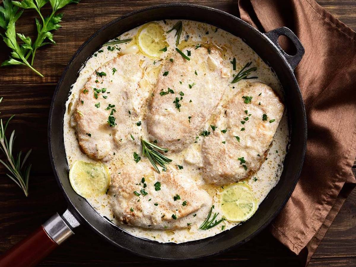 Chicken breasts in creamy sauce with lemon slices and herbs in a skillet on a wooden table.