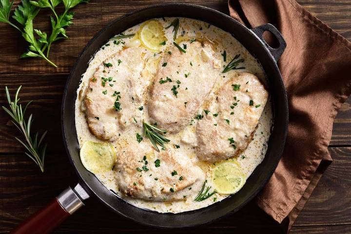 Chicken breasts in creamy sauce with lemon slices and herbs in a skillet on a wooden table.