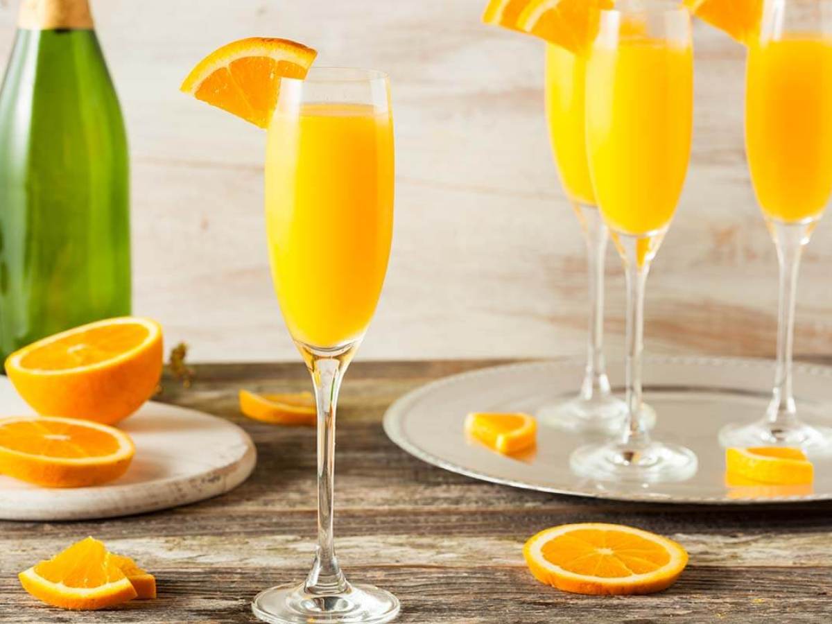 Four mimosa glasses with orange slices on a wooden table, champagne bottle, and orange halves.
