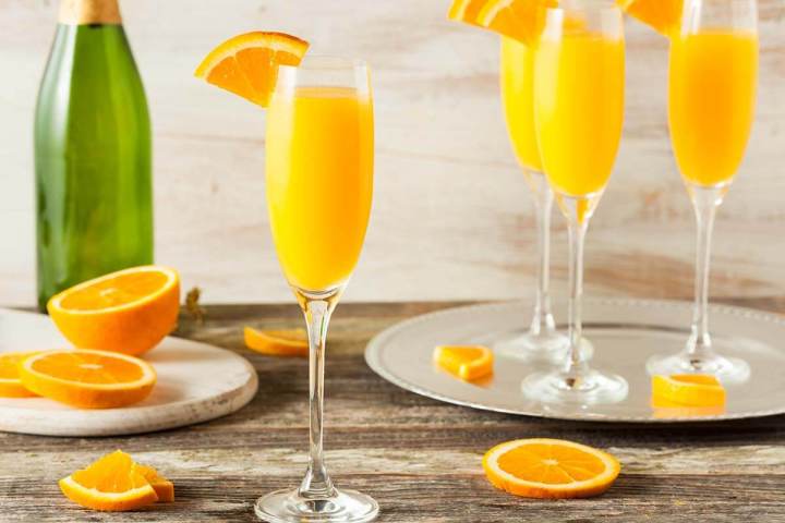 Four mimosa glasses with orange slices on a wooden table, champagne bottle, and orange halves.